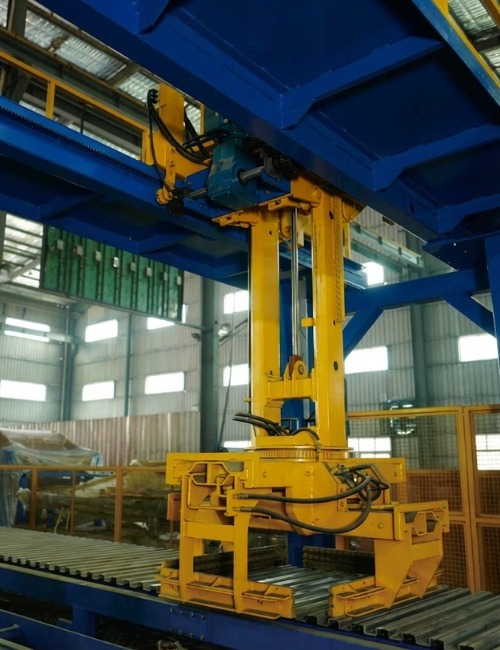 Semi-Automatic Concrete Block Line Hydraulic Handling Unit Close-up of a hydraulic lifting and handling mechanism on a semi-automatic concrete block production line, mounted above a roller conveyor inside an industrial factory setting.